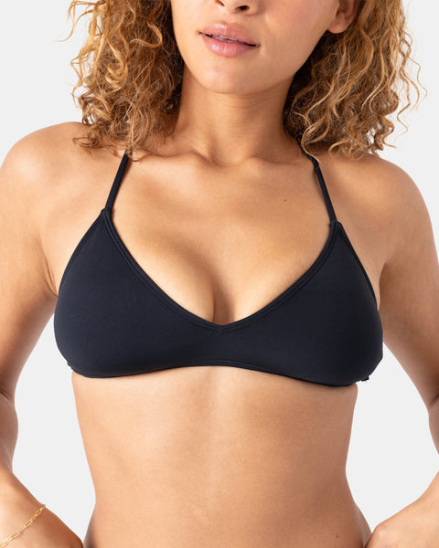 A woman with curly hair models the Roxy Sd Beach Classics Athletic Tri—an athletic black bikini top made from recycled nylon with thin straps, shown from shoulders to midriff against a neutral background.