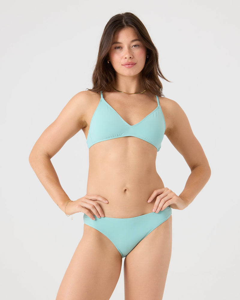 A woman with long brown hair poses against a plain background, wearing a light blue Roxy SD Beach Hipster Bottom Moderate Bikini Bottom. She smiles slightly at the camera with her hands on her hips.