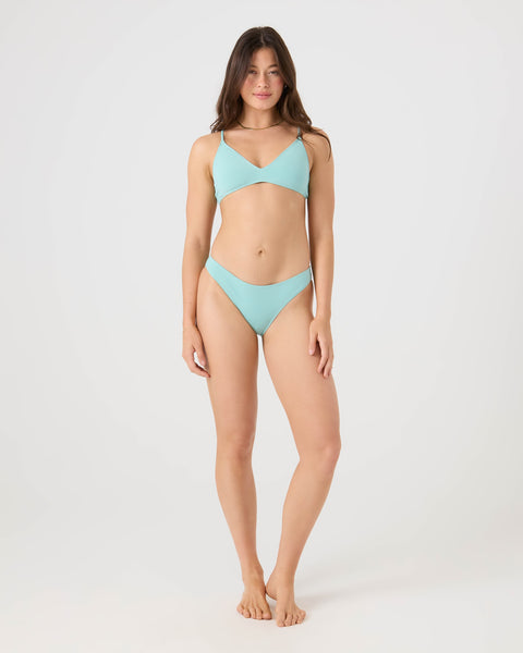 A woman with long brown hair stands barefoot against a white background, wearing the Roxy SD Beach Hipster Bottom Moderate Bikini Bottom. She faces the camera with a relaxed expression, arms resting naturally by her sides.