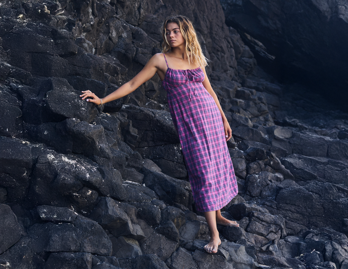 A woman wearing a purple and pink plaid midi dress stands on dark volcanic rocks, holding onto a rock wall.