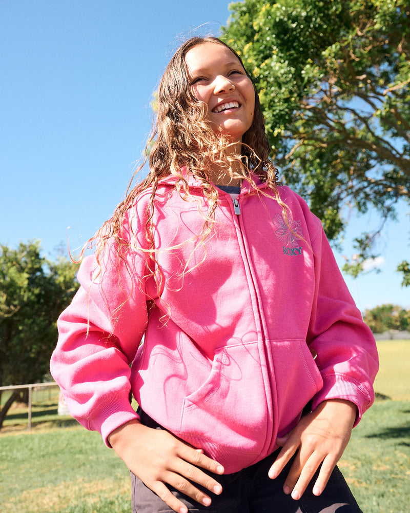 A smiling young person with long, wavy hair wears the Roxy Girls' Hibiscus Stitch Zip Hoodie in hot pink, standing outdoors on a sunny day with trees and blue sky in the background.