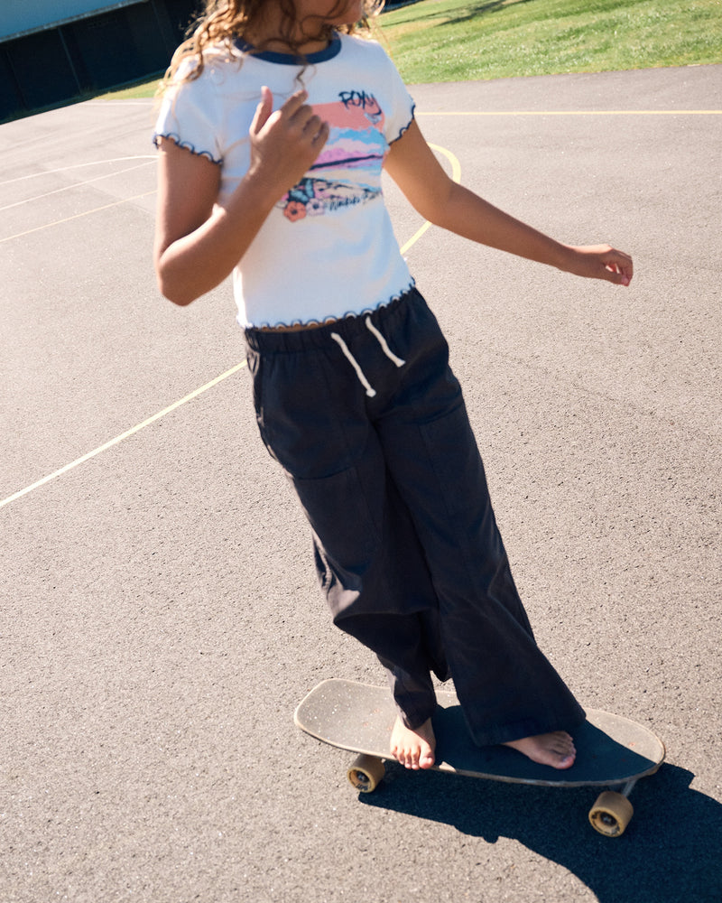 A person with long hair, wearing a white t-shirt and Roxy Girls' Twill Precious Cargo Pant in Phantom, rides a skateboard barefoot on an outdoor paved surface in daylight.