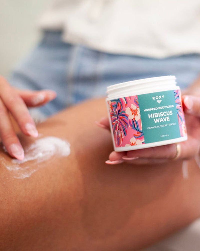 A person applies ROXY’s Hibiscus Wave Body Butter & Scrub Bundle to their leg. The container displays a vibrant floral design, and the product exfoliates, hydrates, and smooths for radiant skin.