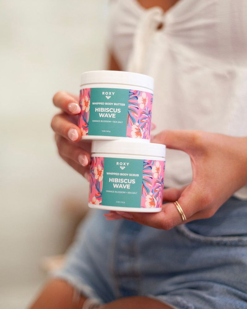 A person in a white top and denim shorts holds two vibrant jars from the ROXY Hibiscus Wave Body Butter & Scrub Bundle, featuring orange blossom and sea salt scents. These luxurious formulas gently exfoliate, hydrate, and smooth your skin to reveal a radiant glow.
