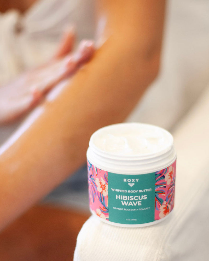 An open jar of ROXY Hibiscus Wave Body Butter & Scrub Bundle sits on a white surface, while someone applies the product to their arm in the background. The jar displays a vibrant floral design and provides exfoliating, hydrating, and smoothing benefits for radiant skin.