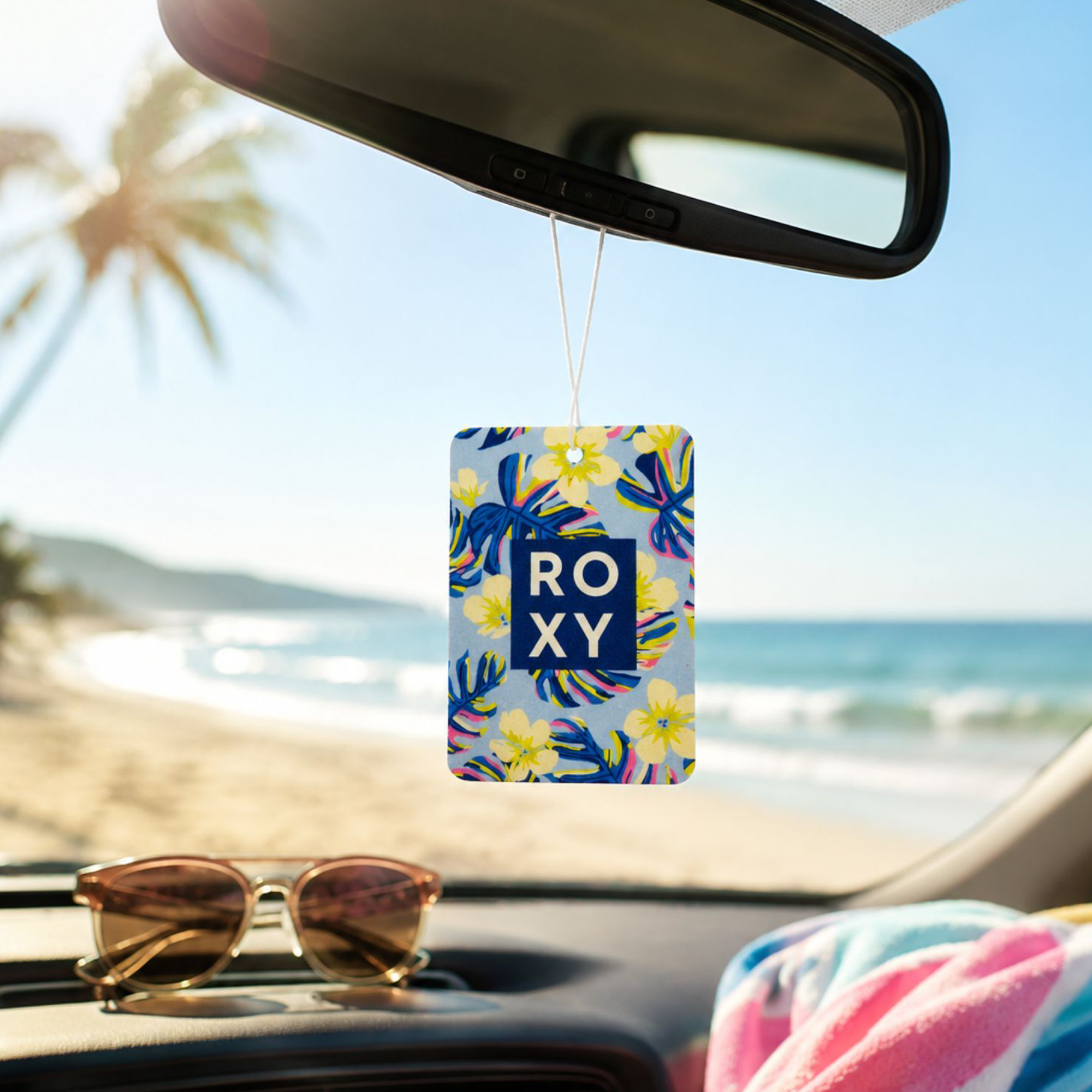 A Deep Dive Palm Air Freshener by Roxy hangs from the rearview mirror. Sunglasses and a towel rest on the dashboard as coconut and ocean air scents float in, with palm trees swaying under a sunny blue sky.