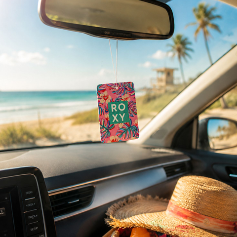 A Roxy Hibiscus Wave Palm Air Freshener with a coastal scent hangs from the rearview mirror, while a straw hat sits on the passenger seat. Outside, palm trees and a sunny beach frame a stunning blue ocean view.
