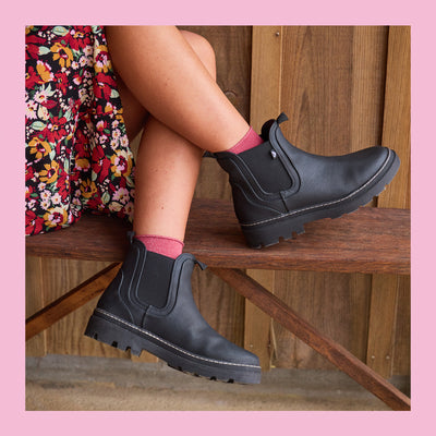 Person wearing black ankle boots with pink socks sitting on a wooden bench.
