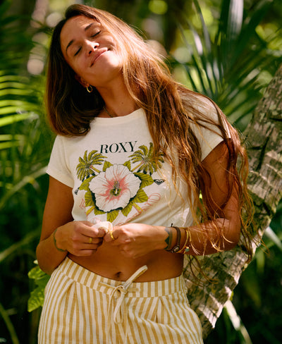 A woman stands outdoors among green tropical plants, eyes closed and smiling softly. She wears a white Roxy t-shirt with a floral design and light yellow striped shorts. Sunlight filters through the foliage.