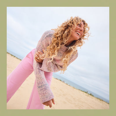 Woman with curly hair wearing a pink sweater and pink pants on a beach.
