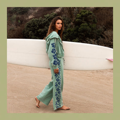 Woman in a green outfit with floral patterns standing next to a white surfboard on a beach.