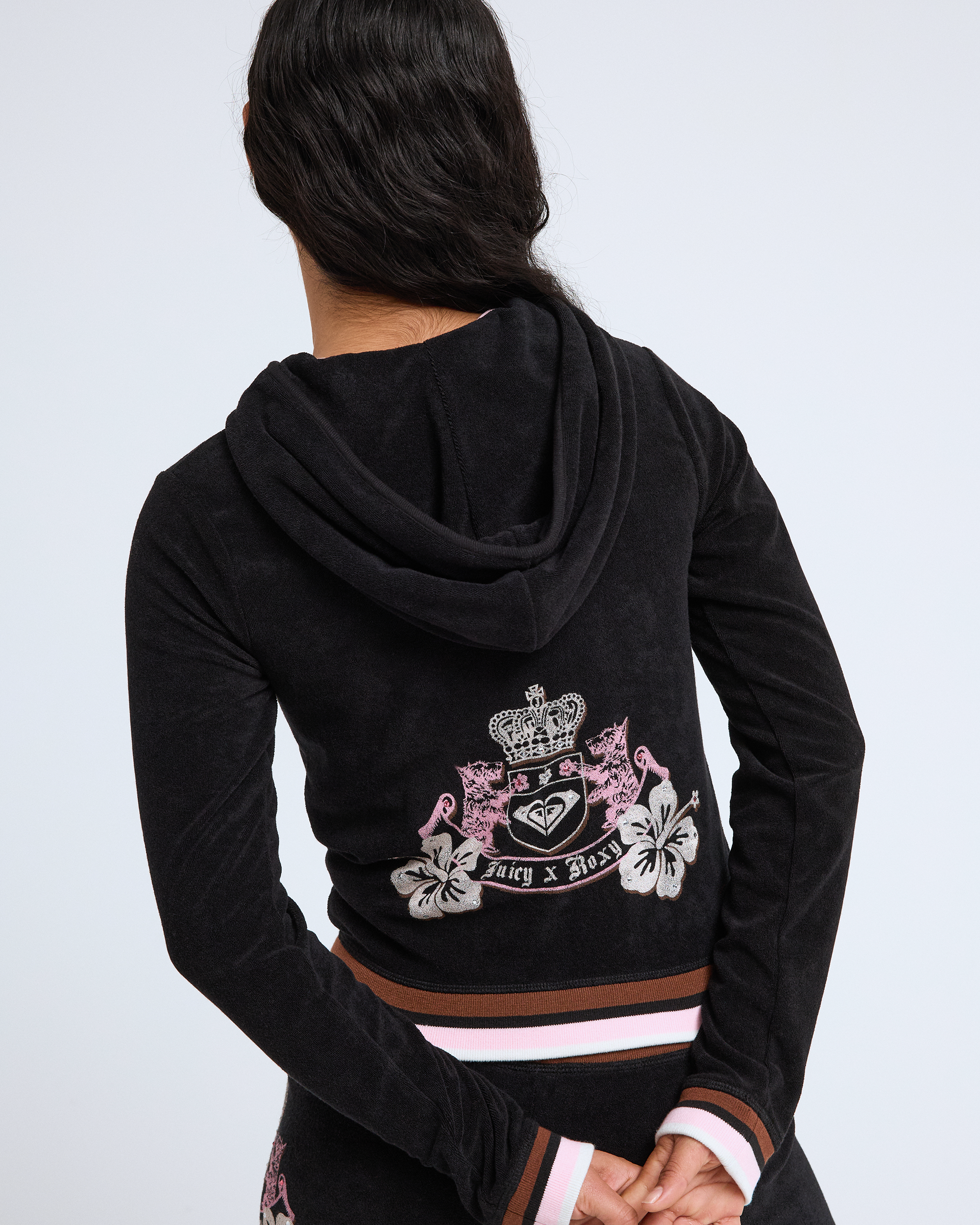 A person with long dark hair, seen from behind, wears a black Roxy x Juicy Fitted Hoodie in Y2K style, featuring a pink, white, and brown striped waistband and an ornate crest design reading “Roxy x Juicy.”.