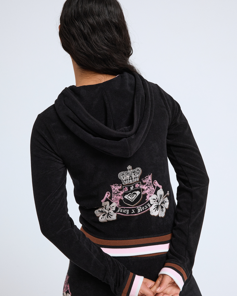 A person with long dark hair, seen from behind, wears a black Roxy x Juicy Fitted Hoodie in Y2K style, featuring a pink, white, and brown striped waistband and an ornate crest design reading “Roxy x Juicy.”.