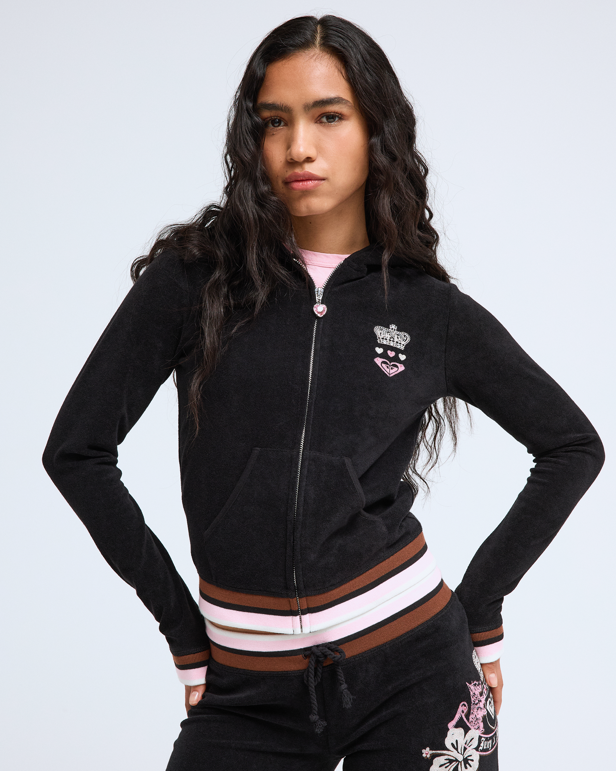 A woman with long wavy hair models the Roxy x Juicy Fitted Hoodie by Roxy, featuring striped cuffs and waistband, with matching pants and decorative patches, evoking Y2K style as she stands hands on hips against a plain light background.
