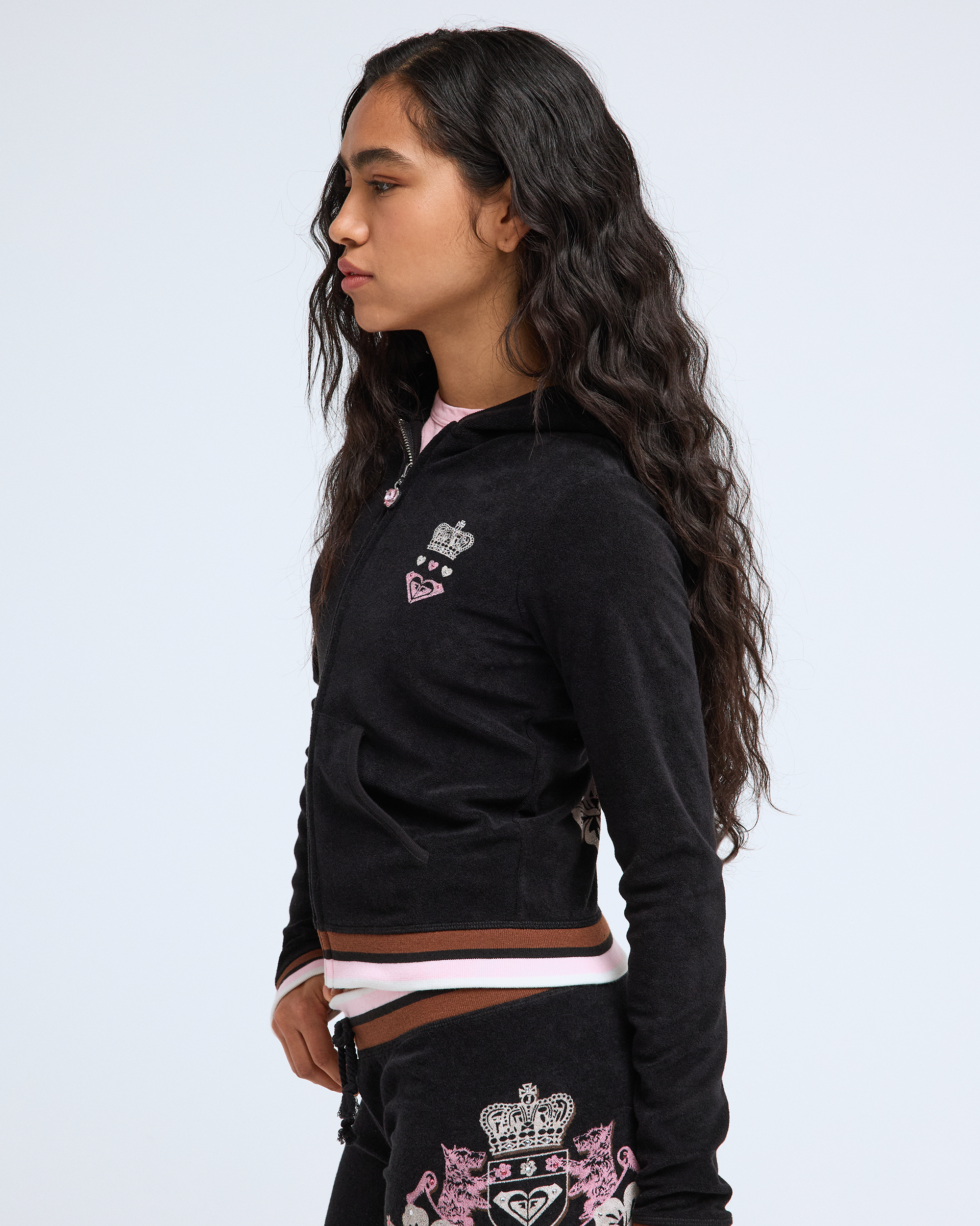 A young woman with long wavy dark hair wears the Roxy x Juicy Fitted Hoodie by Roxy, featuring embroidered crown and heart motifs for a Y2K vibe, standing in profile against a plain white background.