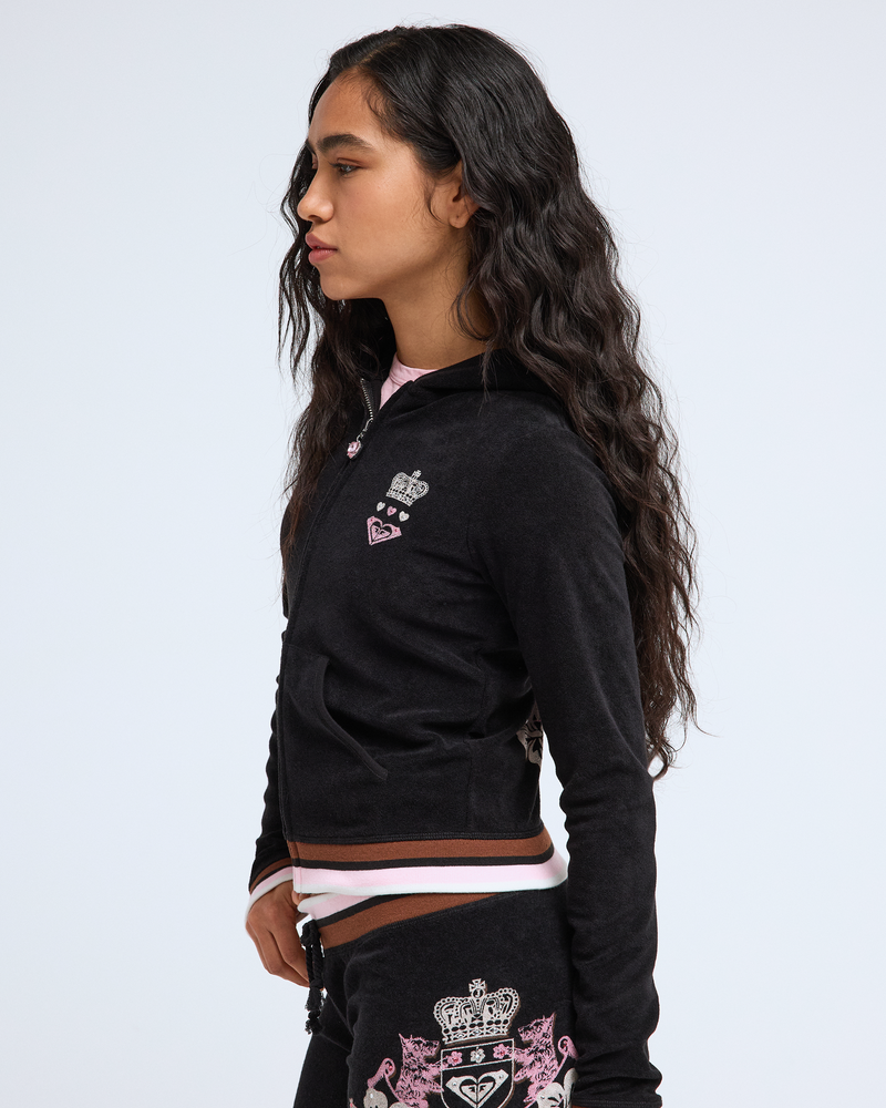 A young woman with long wavy dark hair wears the Roxy x Juicy Fitted Hoodie by Roxy, featuring embroidered crown and heart motifs for a Y2K vibe, standing in profile against a plain white background.