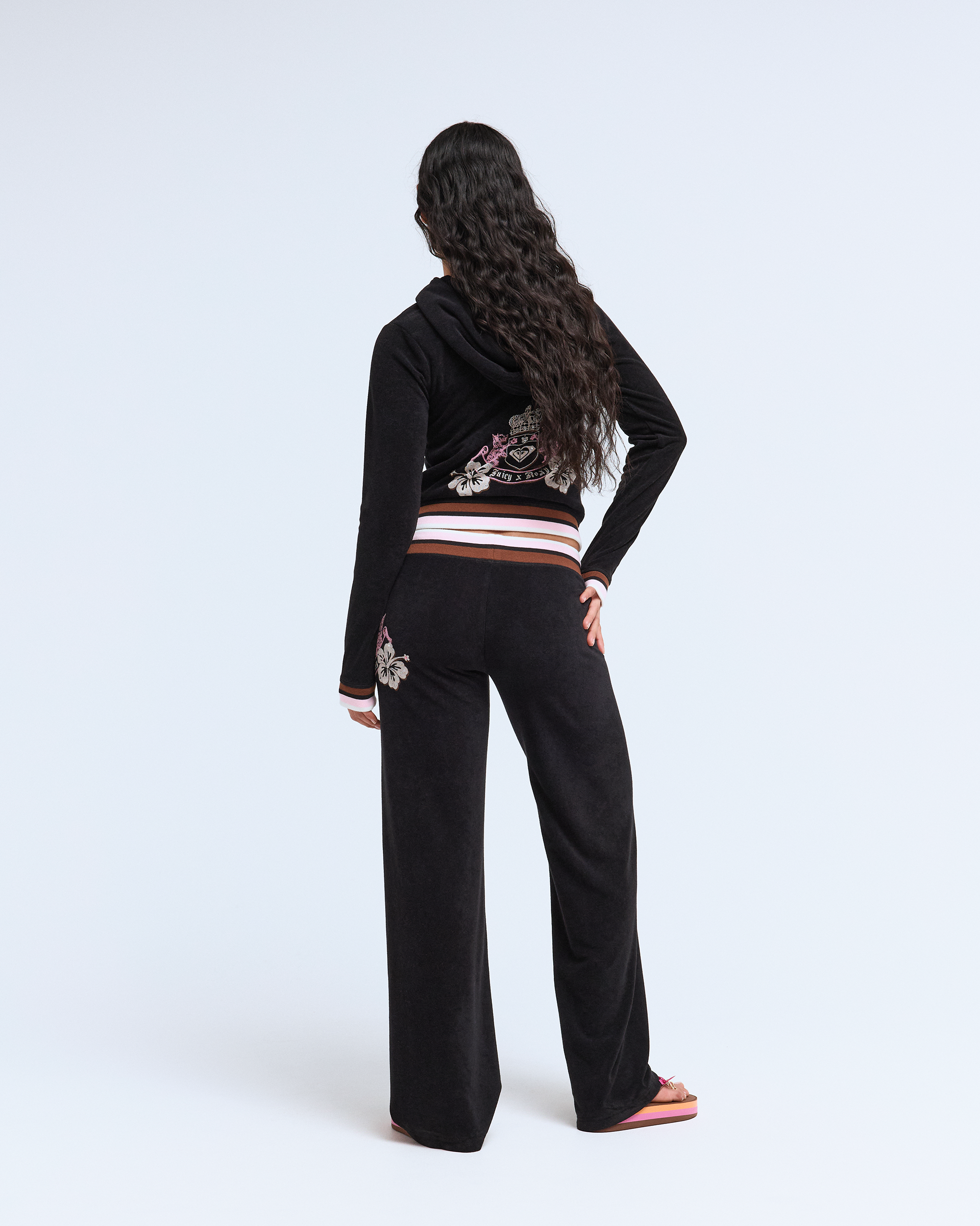 A person with long, wavy dark hair faces away in a black hooded tracksuit and Roxy x Juicy OG Straight Pants by Roxy, featuring floral embroidery in Y2K style, paired with pink flip-flops, set against a plain white background.