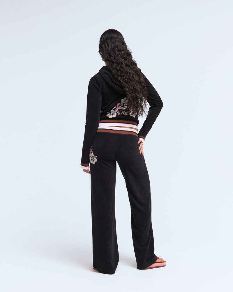 A person with long, wavy dark hair faces away in a black hooded tracksuit and Roxy x Juicy OG Straight Pants by Roxy, featuring floral embroidery in Y2K style, paired with pink flip-flops, set against a plain white background.