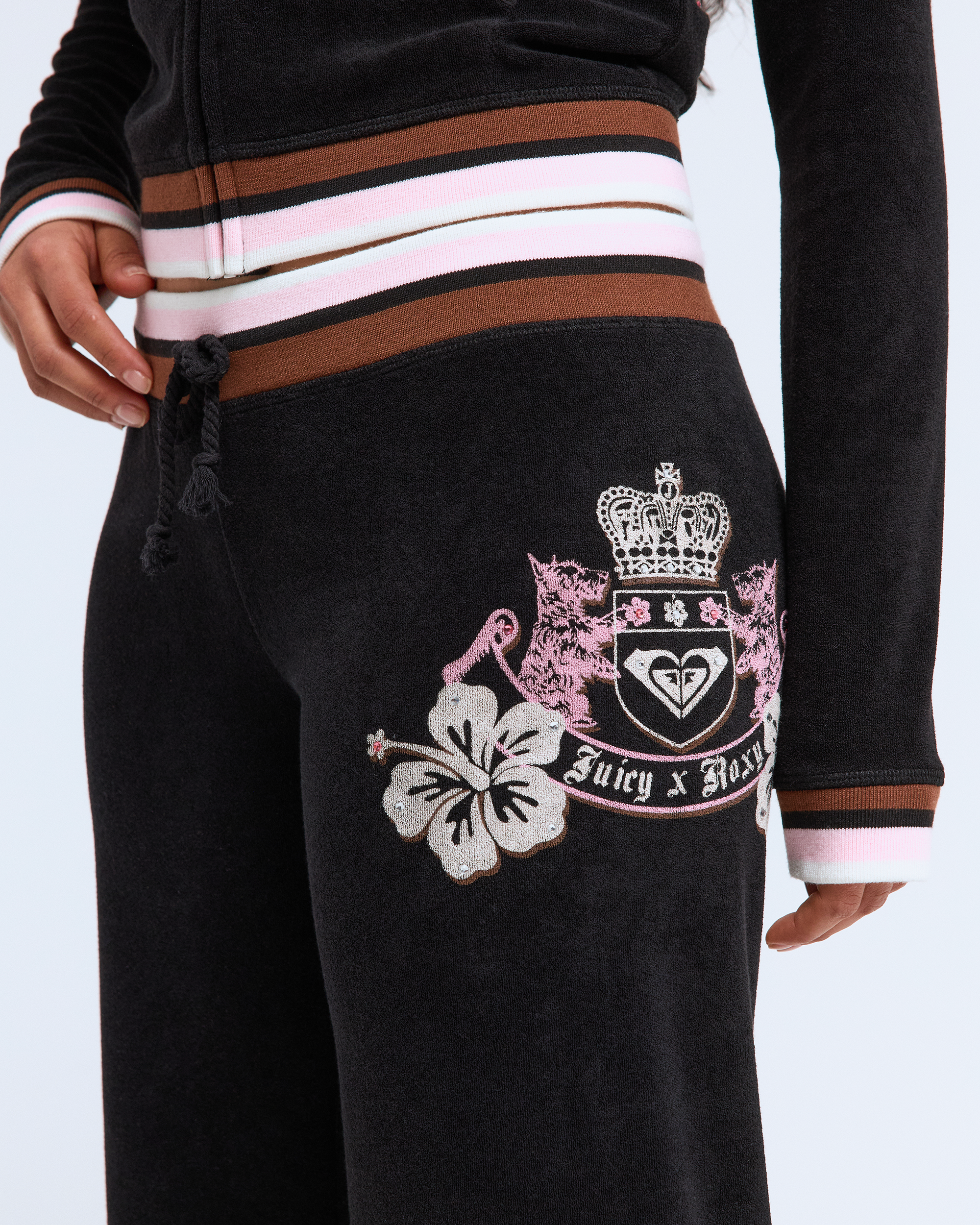Wearing Roxy x Juicy OG Straight Pants in black velour with a floral and crest emblem at the hip, paired with a matching zip-up jacket featuring pink, white, and brown striped trim for a fresh Y2K-inspired look.