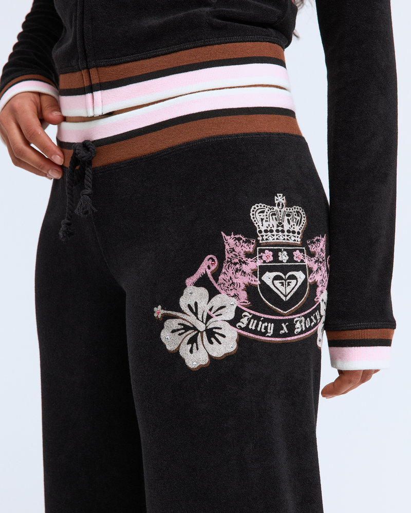 Wearing Roxy x Juicy OG Straight Pants in black velour with a floral and crest emblem at the hip, paired with a matching zip-up jacket featuring pink, white, and brown striped trim for a fresh Y2K-inspired look.