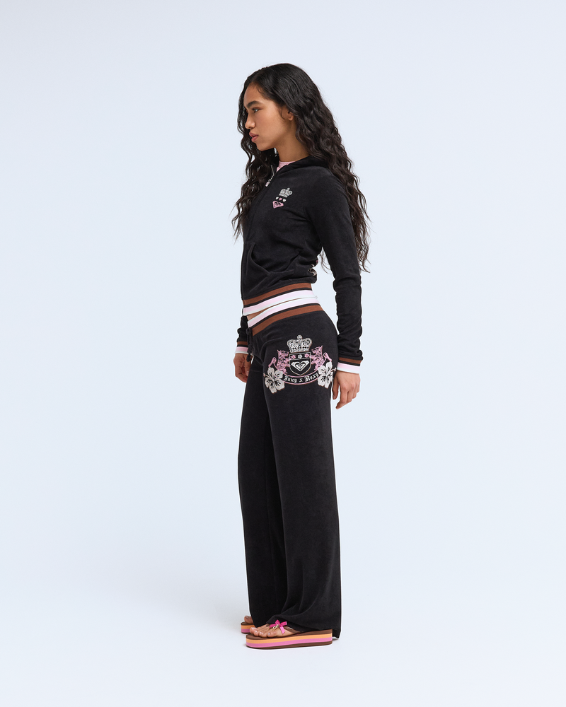 A woman with long, wavy hair stands sideways in Y2K style, wearing Roxy x Juicy OG Straight Pants and a black tracksuit with embroidered details, paired with pink platform sandals against a plain white background.