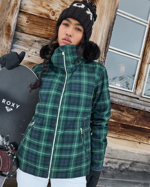 A woman in a green plaid Roxy Jet Ski Jacket with cozy insulation and a knit hat stands outside a wooden cabin holding a snowboard. Snow and mountains are visible in the background, highlighting her Jet Ski Jacket’s waterproof protection against the winter elements.
