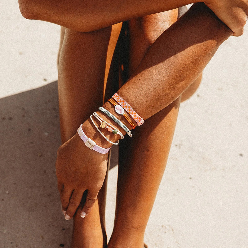 A sun-kissed person sits cross-legged on sandy ground, radiating relaxed beach vibes, adorned with colorful K'lani x Roxy Hair Tie Bracelets from Roxy on their wrists and ankles.