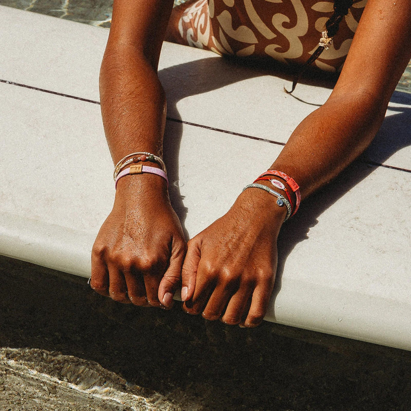 A person with tan skin in a patterned Roxy Pink Tropics swimsuit rests at the pool's edge, sunlight highlighting colorful K'lani x Roxy Hair Tie Bracelets by Roxy on their wrists along with natural stone beads.