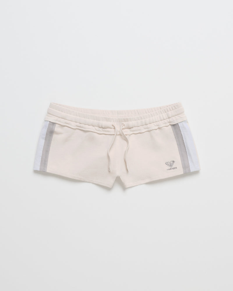 The ROXY x Madhappy French Terry Mini Short features a beige colorway with an elastic waistband, drawstring, and gray side stripes, displayed flat on a white background. The bottom left leg is detailed with a small logo that highlights the Roxy x Madhappy collaboration.