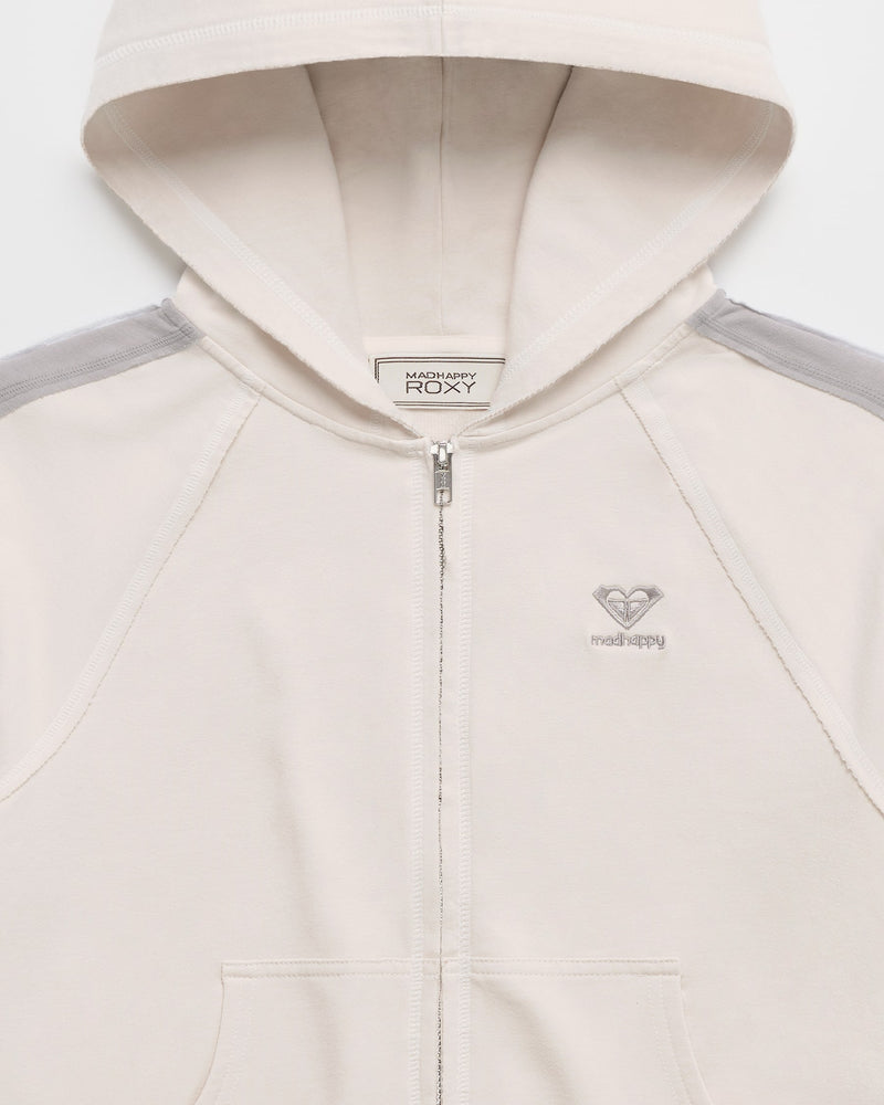 Introducing the ROXY x Madhappy French Terry Shrunken Full Zip Jacket—a limited-edition beige hoodie crafted from soft French Terry, featuring gray shoulder accents, two front pockets, and a small embroidered logo on the chest. The hood is worn up with a white clothing label visible inside the neckline.