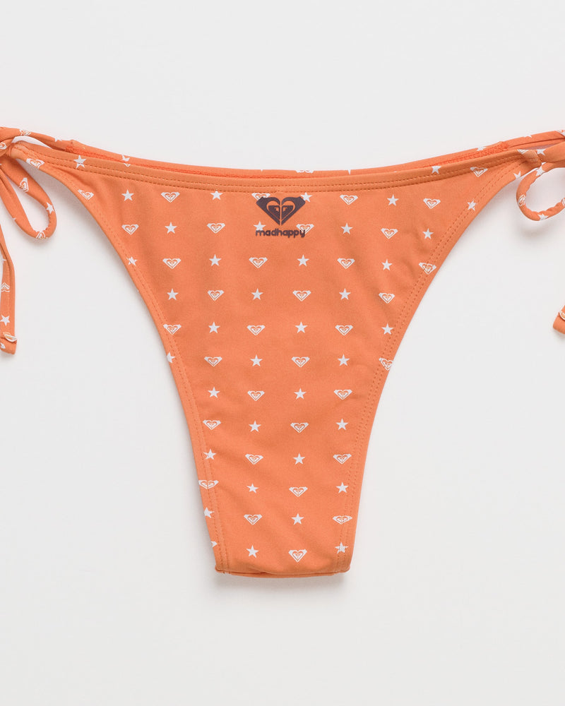 The ROXY x Madhappy Tie Side Cheeky Bikini Bottom features a vibrant orange hue adorned with white star and diamond patterns. Designed with adjustable side tie strings and a small MadHappy logo at the center front, this playful piece from the Roxy x Madhappy collaboration is shown laid flat against a white background.