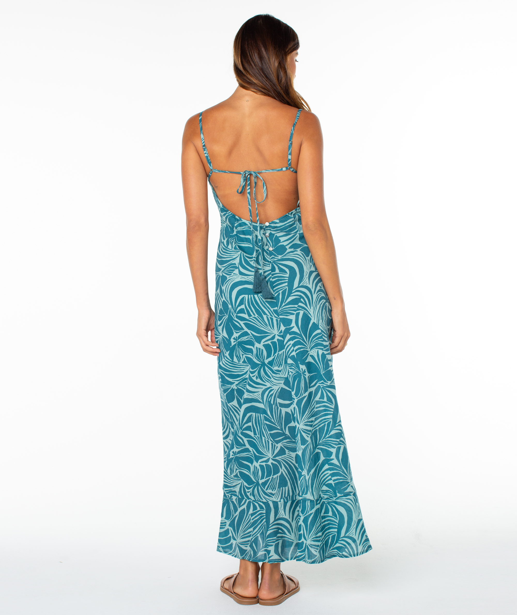 A woman with long brown hair wears the Roxy Lost Paradise Dress, a teal maxi dress with a white leaf print and an open, strappy back. She pairs it with sandals and stands on a white background, facing away from the camera.