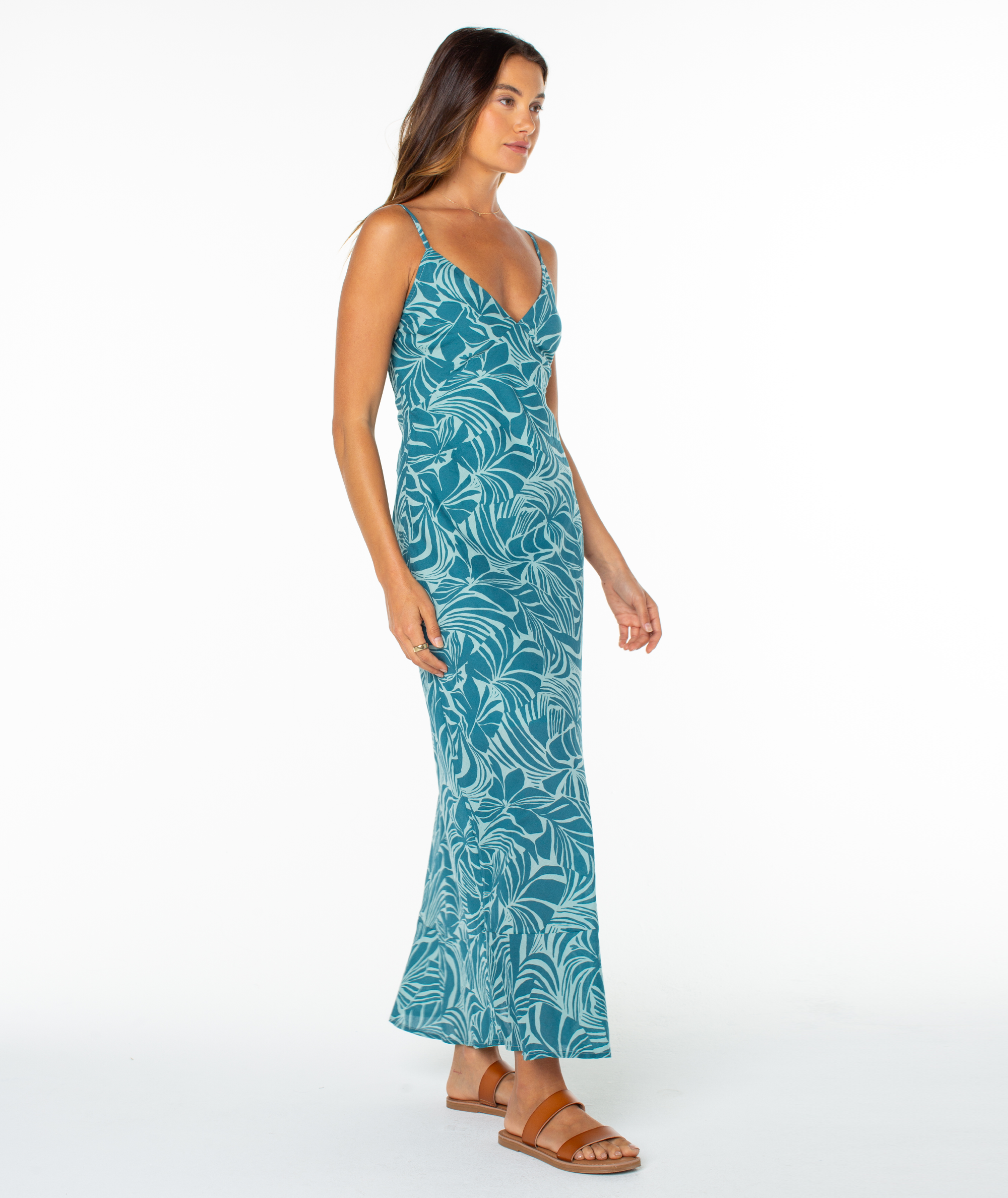 A woman models the Roxy Lost Paradise Dress—a teal, sleeveless, ankle-length maxi with a leaf print and open back—paired with sandals against a plain white background, gazing slightly to the side.