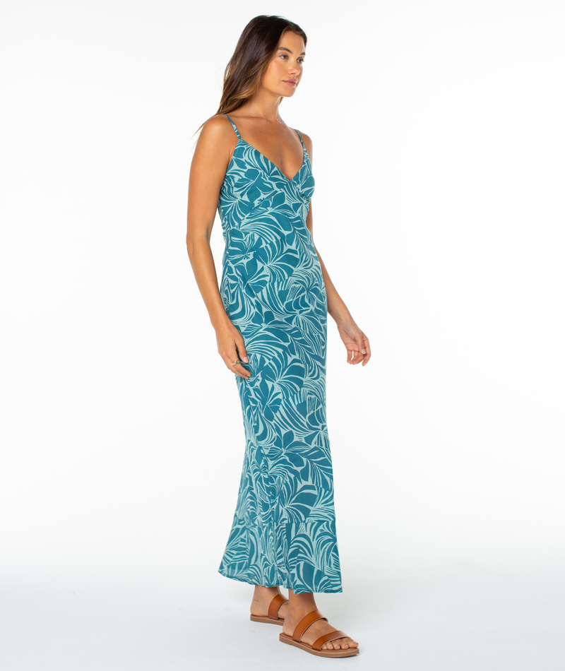 A woman models the Roxy Lost Paradise Dress—a teal, sleeveless, ankle-length maxi with a leaf print and open back—paired with sandals against a plain white background, gazing slightly to the side.