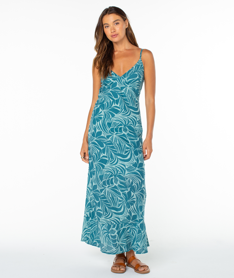 A woman with long, wavy hair poses casually against a white background in the Roxy Lost Paradise Dress, which features a leafy print and open back. She completes the look with brown sandals.