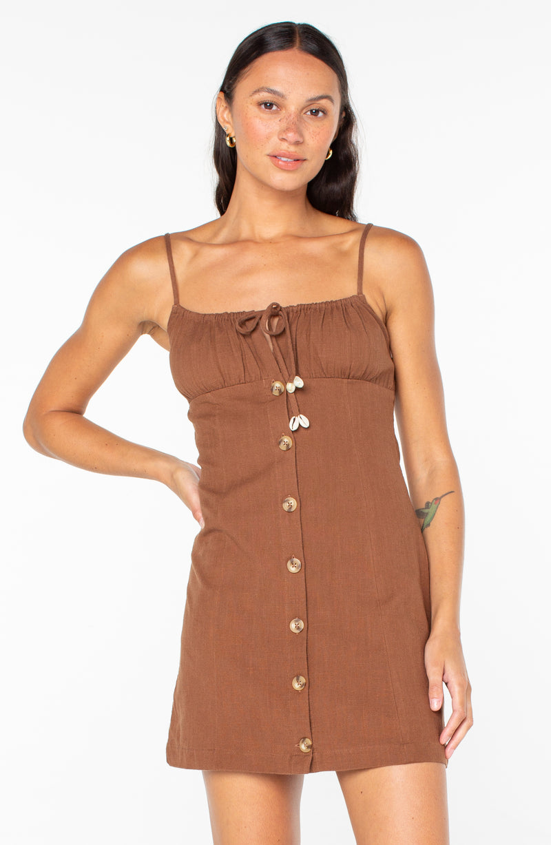 A woman wears the Roxy Somewhere Sunny mini dress in Tiramisu Solid, featuring spaghetti straps, a tie-front bodice, and a button-down front with seashell accents.