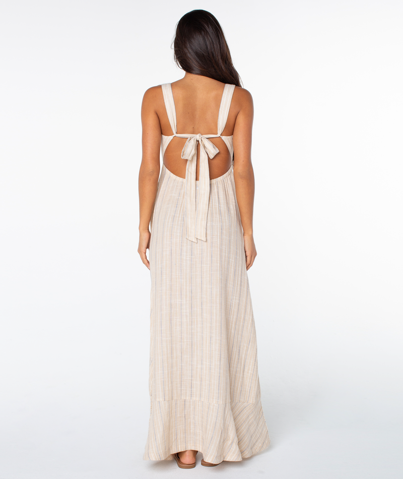 A woman faces away in the Roxy Wilder Coast Dress—a long, sleeveless light beige maxi with subtle stripes, wide open-back straps, a large bow at mid-back, and an elegant V neckline, all set against a plain white background.