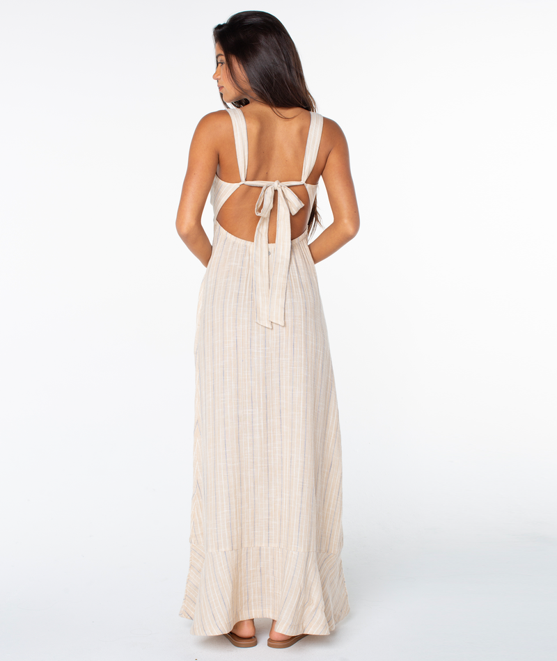 A woman with long dark hair stands with her back to the camera, wearing the Roxy Wilder Coast Dress— a light beige maxi dress featuring thin vertical stripes, an open tie-back bow, and a subtle V-neckline—against a white background.