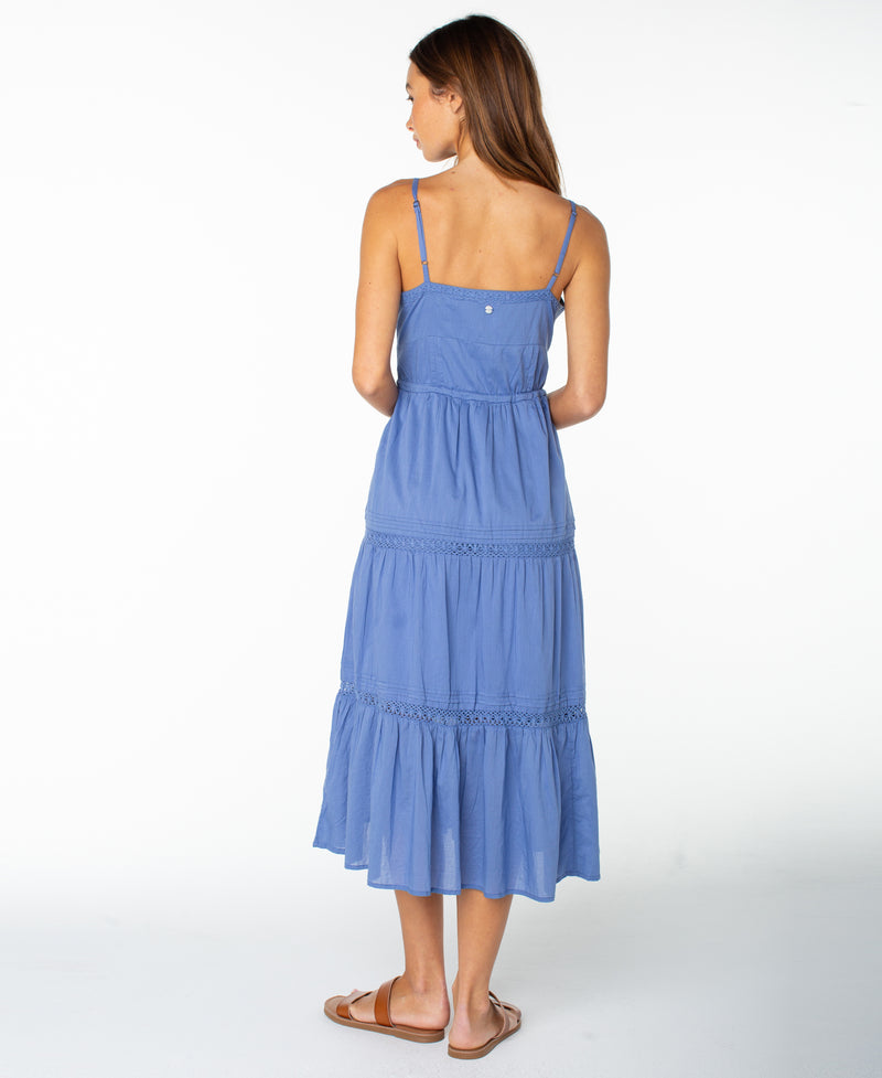 A woman stands facing away, wearing the sleeveless blue Azalea Dress by Roxy—a tiered midi dress perfect for transitional seasons—paired with brown sandals, set against a plain white background.