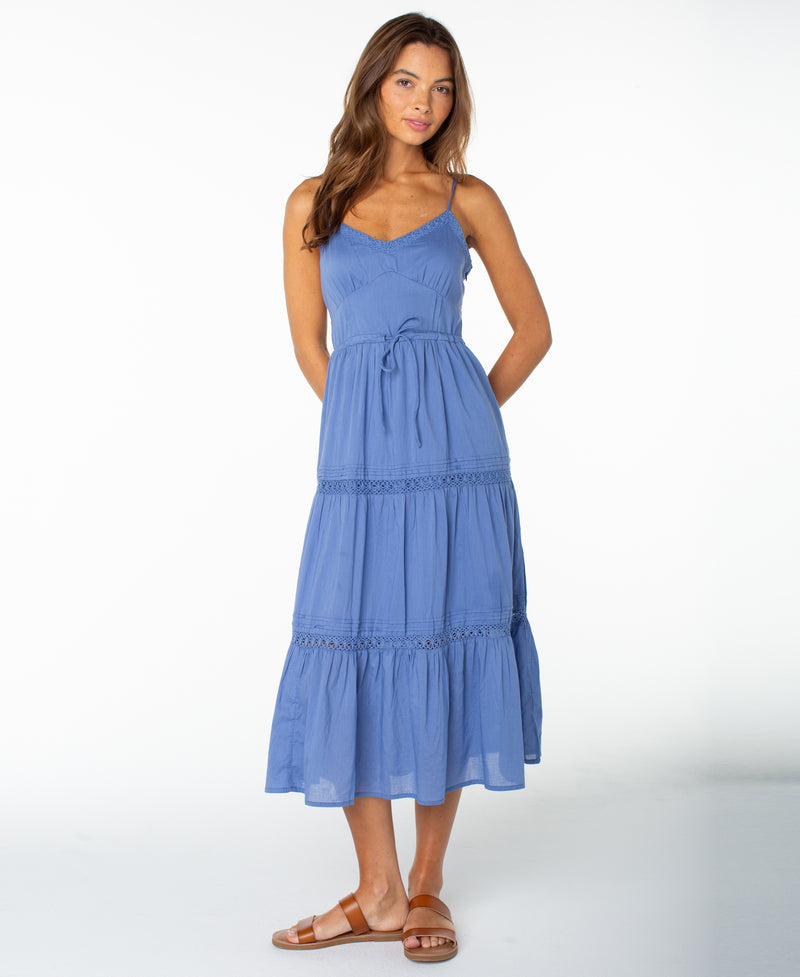 A woman models the Roxy Azalea Dress in Dutch Blue Solid, a sleeveless style featuring a waist tie, paired with brown sandals and set against a plain white background. This versatile dress is perfect for transitioning between seasons.
