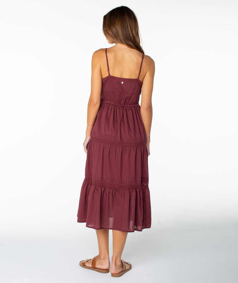 A woman with long brown hair stands with her back to the camera, wearing the sleeveless maroon Azalea Dress by Roxy—a tiered midi dress ideal for transitional seasons—paired with brown sandals against a plain white background.