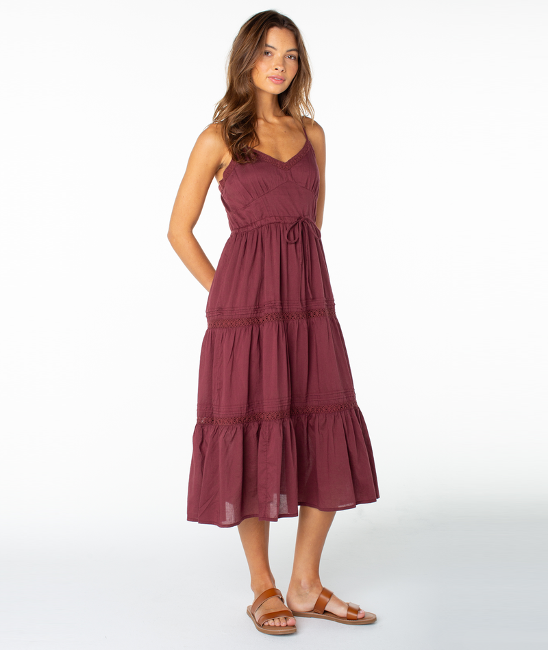 A woman stands wearing the Roxy Azalea Dress—a sleeveless, maroon, tiered midi dress—paired with sandals. Her long, wavy hair frames her face as she places her hands behind her back. Perfect as a transitional piece, this look stands out against the plain white background.