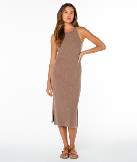 A woman with long wavy hair poses against a white background in the Roxy Emilia Midi Dress, a sleeveless, fitted, ribbed taupe cotton blend style. She pairs it with brown flat sandals and stands relaxed with one hand on her hip.