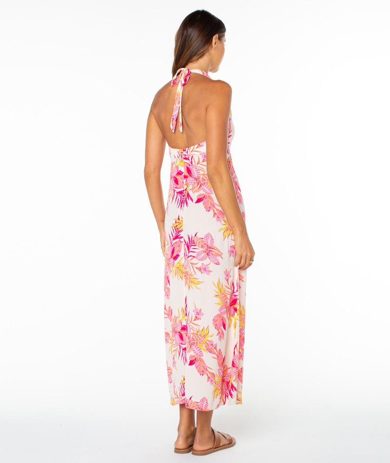 A woman stands facing away wearing the Roxy Beach Day Maxi Dress, featuring a halter neckline, backless ankle-length design with pink, yellow, and orange florals, paired with brown sandals against a white background.