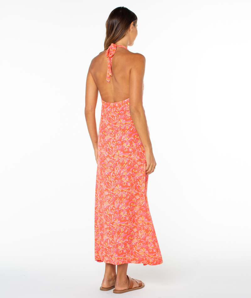A woman faces away in the Roxy Beach Day Maxi Dress, featuring a halter neckline and bright pink and orange floral print. She has long hair, wears flat sandals, and stands against a plain white background.