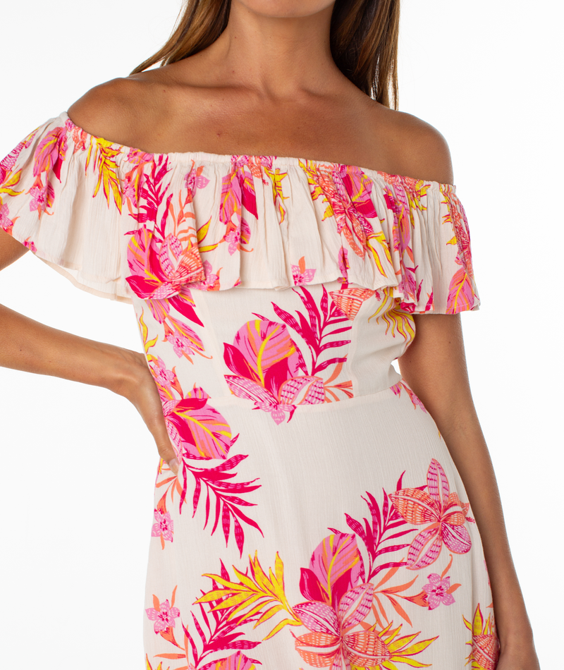 A woman models the Roxy Clear Sky Mini Dress, a white, off-shoulder mini dress with ruffles and pink, orange, and yellow tropical floral patterns. The image is cropped at her shoulders and above the knees, with her left hand on her hip.