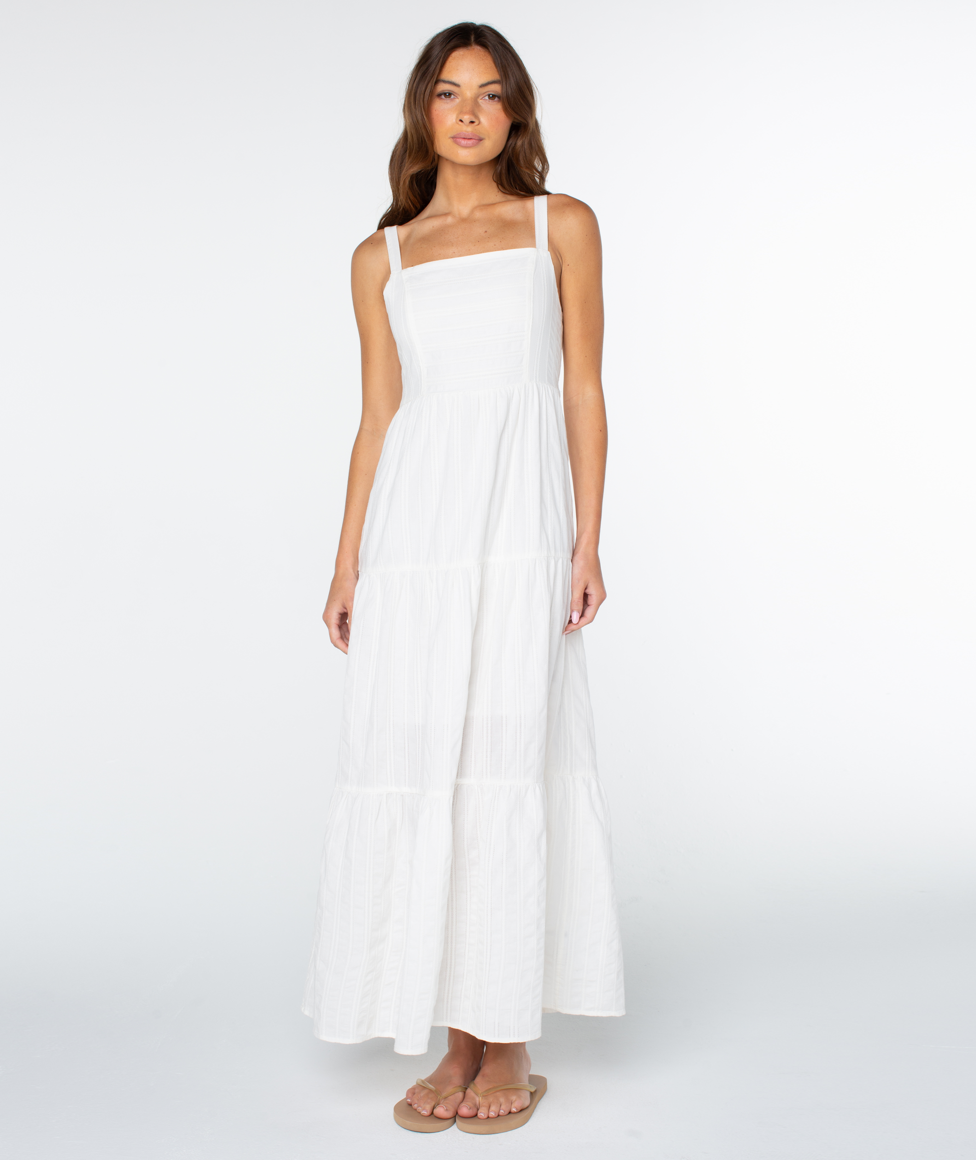 A woman with long brown hair wears the Roxy Clearwater Maxi Dress, a sleeveless white ankle-length vacation dress with a square neckline and tiered skirt. She stands facing forward on a plain white background in beige sandals.