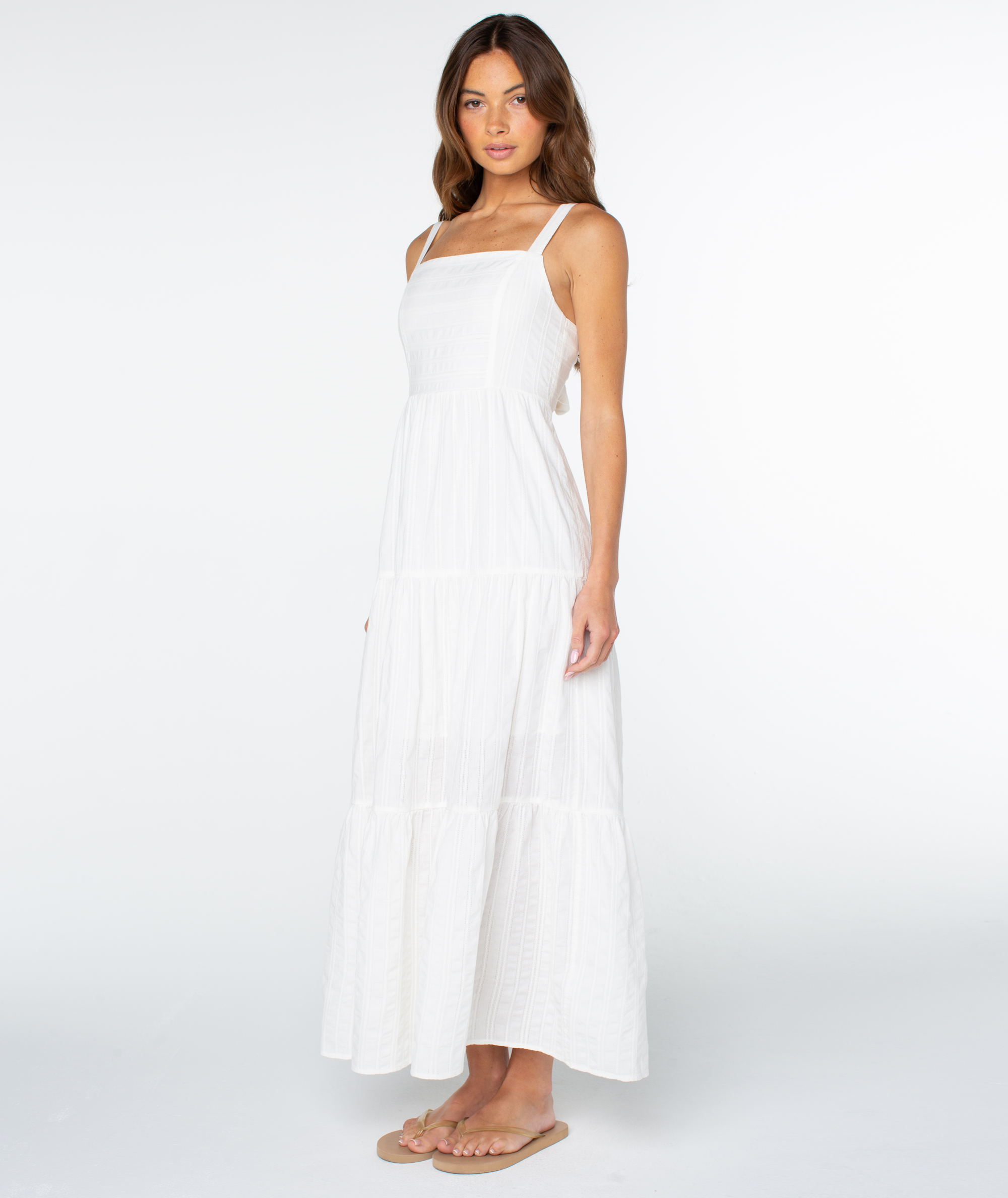 A woman with long brown hair and a neutral expression models the sleeveless, white Roxy Clearwater Maxi Dress against a plain white background, paired with beige flip-flops.