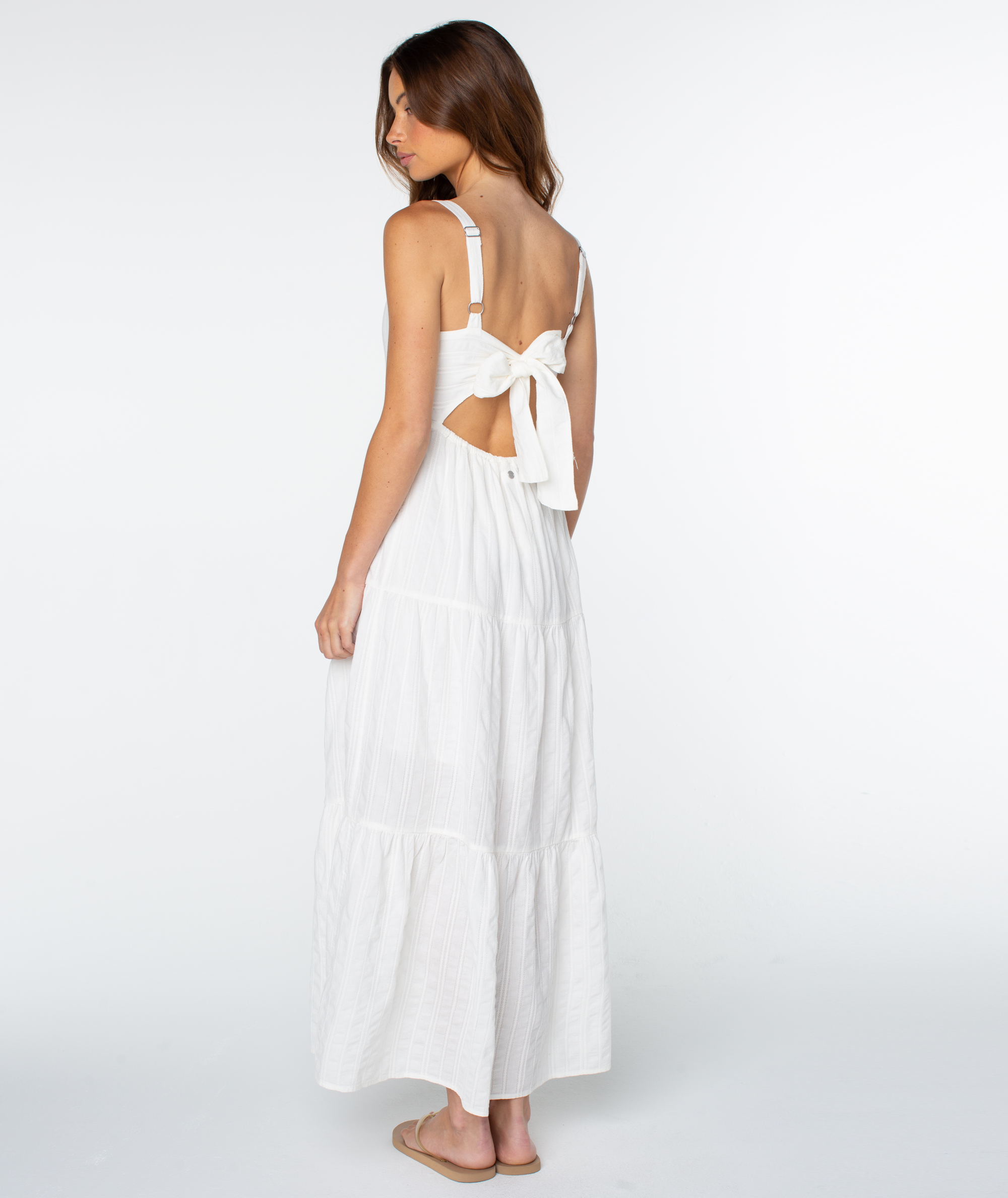 Model seen from behind, wearing the Roxy Clearwater Maxi Dress—a white tiered vacation dress featuring a bow and cutout detail on the back.