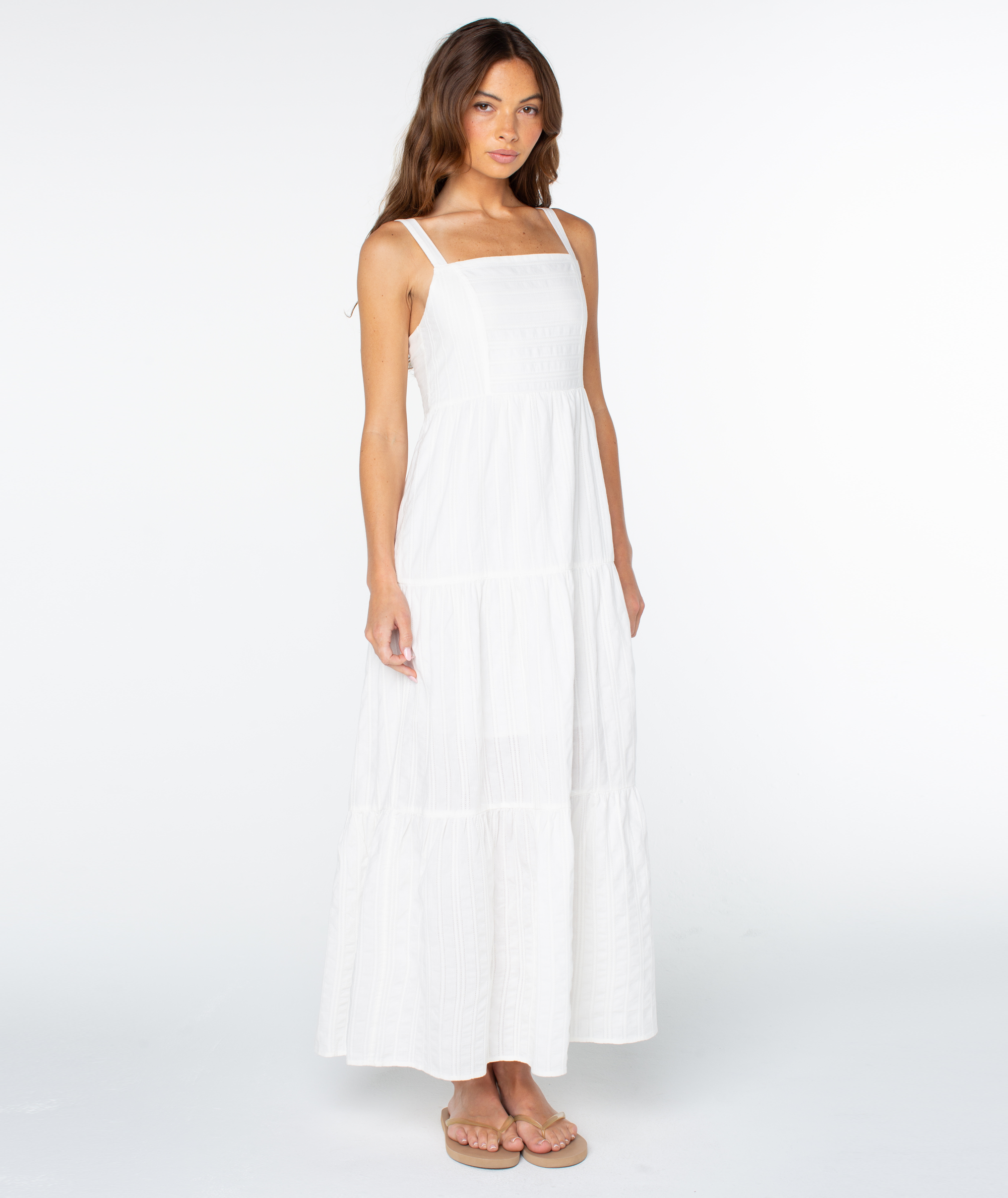 A woman with long brown hair wears the Roxy Clearwater Maxi Dress, featuring a sleeveless, white, square-neck design and beige sandals, standing against a plain white background.
